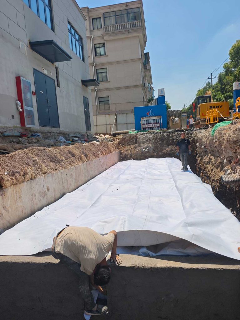 建筑旁基坑内,工人们正在对覆盖了白色防渗土工布的小型硅砂雨水收集池进行表面铺设与细节调整