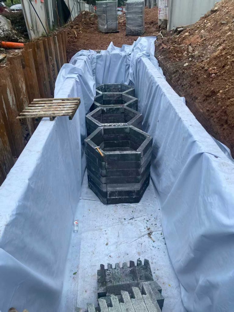 铺有白色防渗土工布的狭长基坑内,中央已拼装完成一排四个正六边形蜂巢状硅砂雨水收集井筒